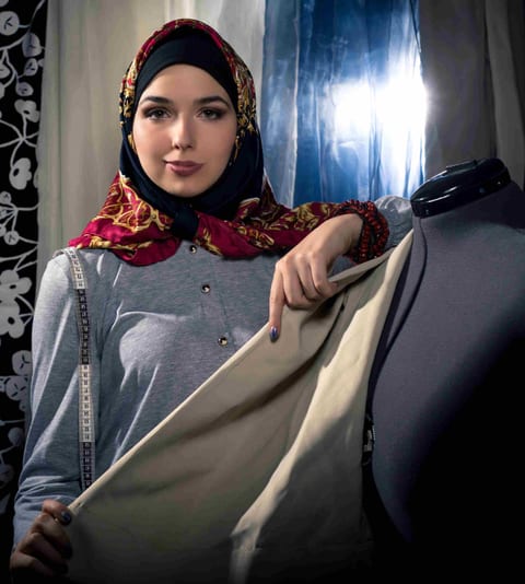 A model in a red-and-gold patterned hijab holds open a cream-lined abaya on a dress form, a tailor's tape measure draped at her side.